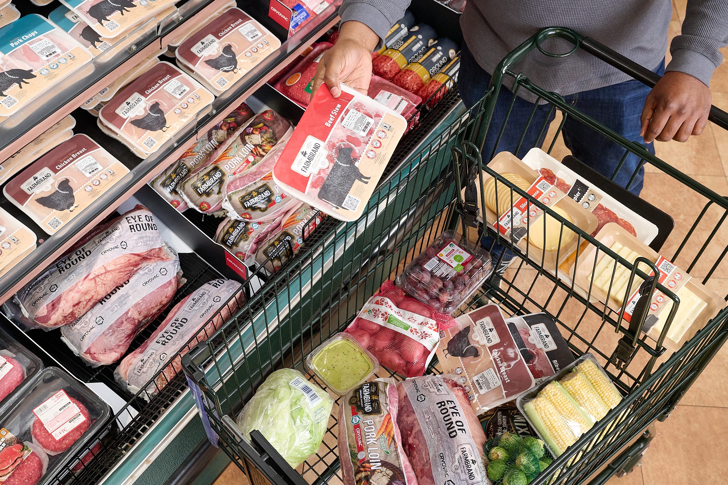 A customer with a full shopping cart is standing in front of a meat case.